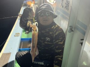 迷彩柄のシャツを着て、サングラスをかけ、帽子をかぶった人物が、夜の漁船でイカをさばいている。背景にはもうひとり、釣りをしている人物が見える。シーンは薄暗い。.