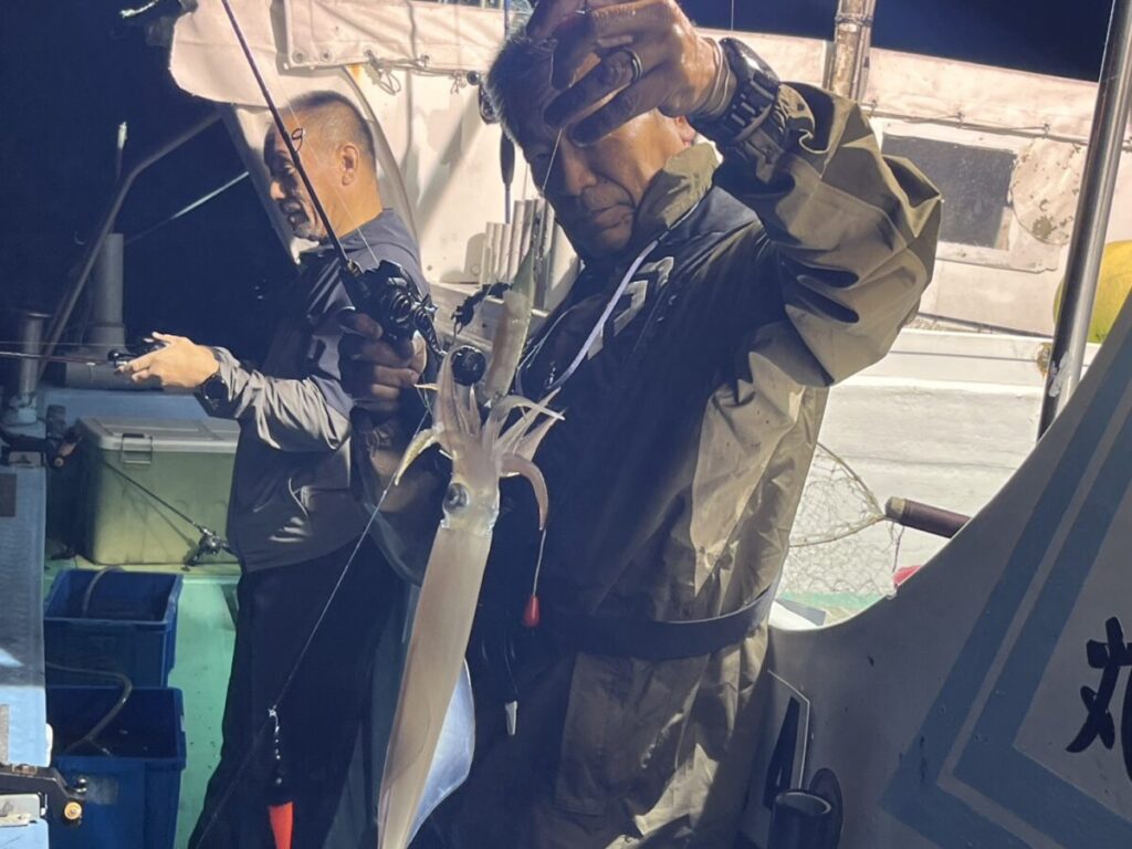 一人は釣り竿を持ち、釣り上げたばかりの大きなイカを見せている。人工的な光に照らされ、イカの体が浮かび上がっている。.