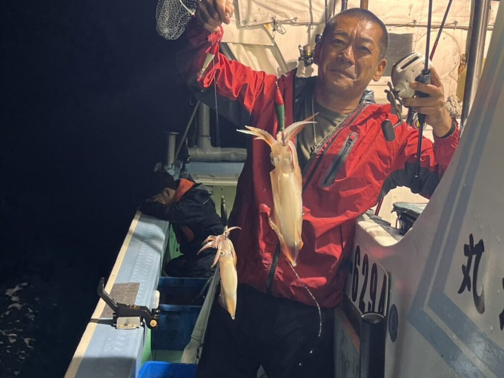 夜、赤いジャケットを着た男がボートの上に立ち、両手にそれぞれ1匹ずつ、獲れたてのイカを誇らしげに掲げている。背景には、姿の一部が見える別の人が座っている。.