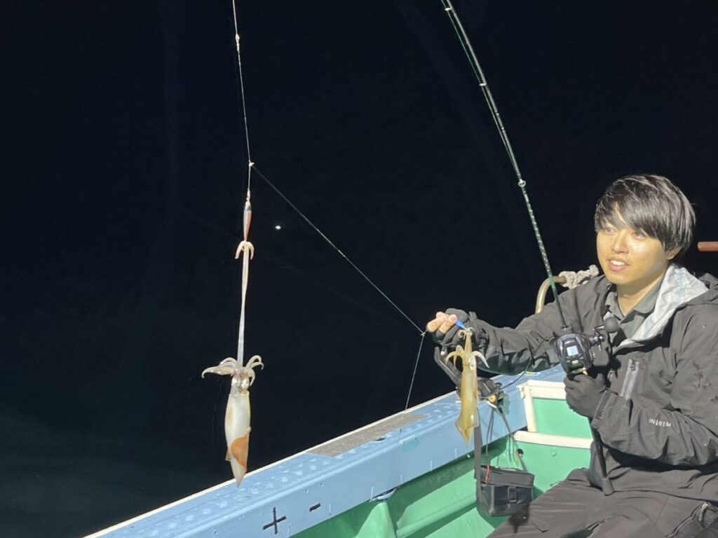 夜、ボートに乗った人物が、釣り竿を手に持ち、その糸には2匹のイカが掛かっており、明るい光に照らされている。.