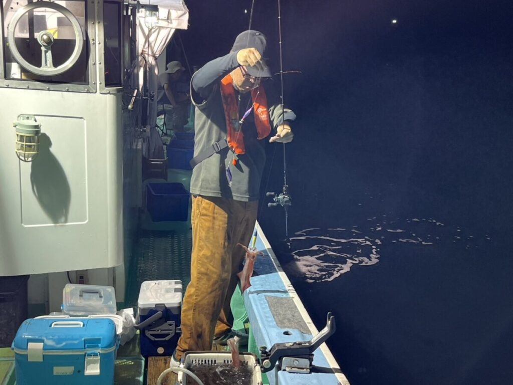 夜、ボートに乗った人物が、人工照明の下で魚を釣り上げている。その人物は帽子、ライフジャケット、茶色のズボンを身に着けており、そばには釣り道具や収納ボックスが置かれている。.