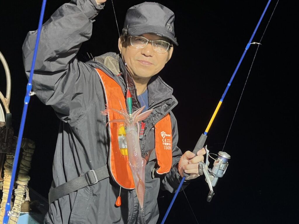 眼鏡をかけ、黒いレインジャケットを羽織り、オレンジ色の救命胴衣を着た人が釣り竿を掲げ、闇夜に釣りたてのイカを見せる。.