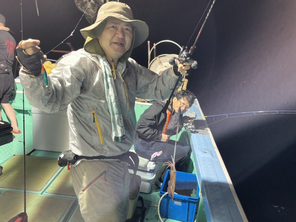 前の人は釣り竿と釣り糸を持って微笑み、後ろの人は青いバケツからイカを巻き上げている。二人ともジャケットと帽子をかぶっている。.