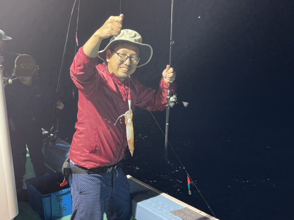 夜の船上で、釣ったばかりのイカを乗せた釣り竿を手に笑顔を見せる赤いジャケットと帽子の人物。.