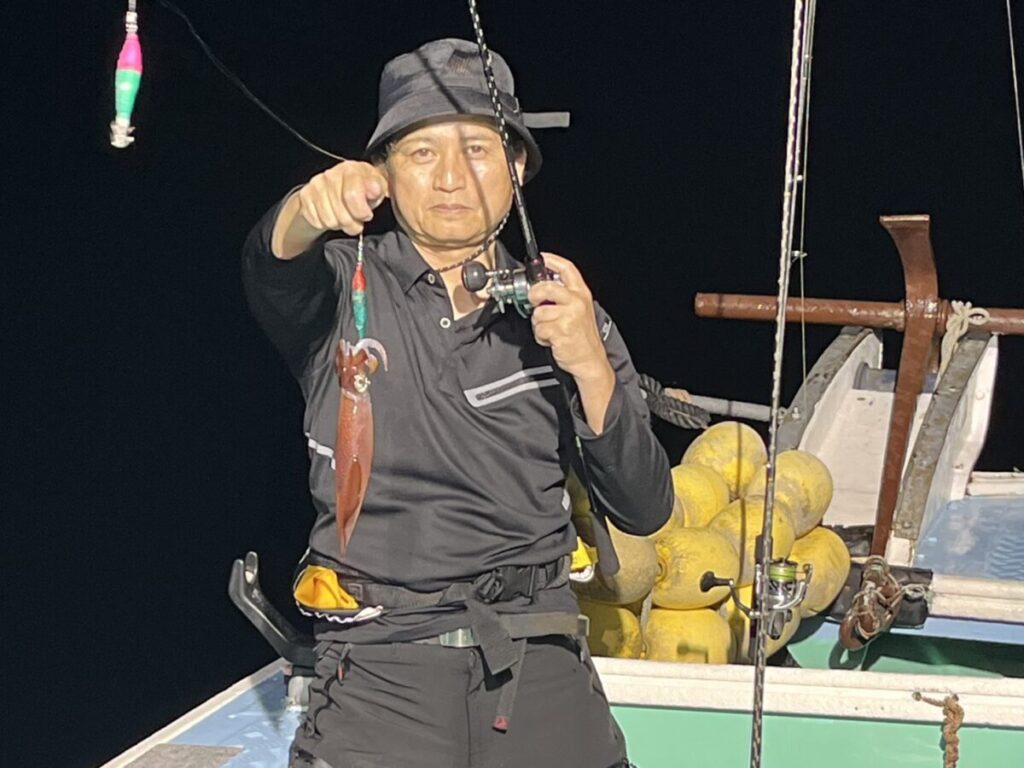 黒い漁具を身につけ、帽子をかぶった人が、夜の船の上で釣り竿を持ち、前方を指さしている。 イカを釣り上げ、釣り糸にぶら下がっている。背後に黄色いブイが見える。.