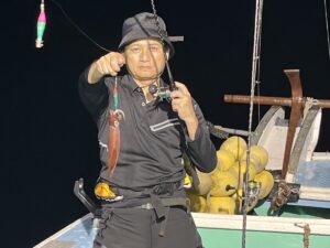 黒い漁具を身につけ、帽子をかぶった人が、夜の船の上で釣り竿を持ち、前方を指さしている。 イカを釣り上げ、釣り糸にぶら下がっている。背後に黄色いブイが見える。.
