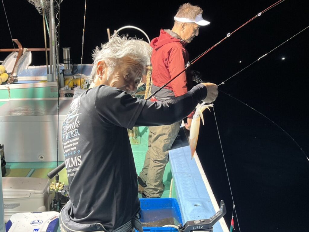 手前の人物は釣ったばかりのイカを笑顔で掲げ、もう一人は船縁近くに立って釣り糸に集中している。.