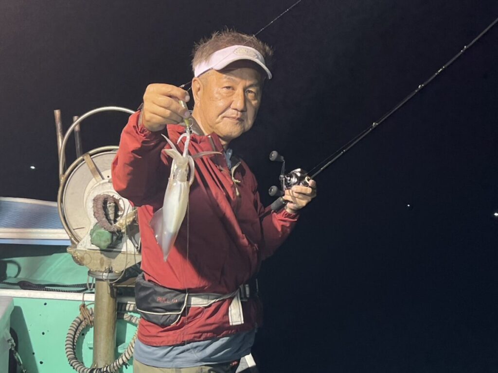 赤いジャケットに白いバイザーの男が、釣り竿を片手に夜の船上で釣りたてのイカを掲げている。.