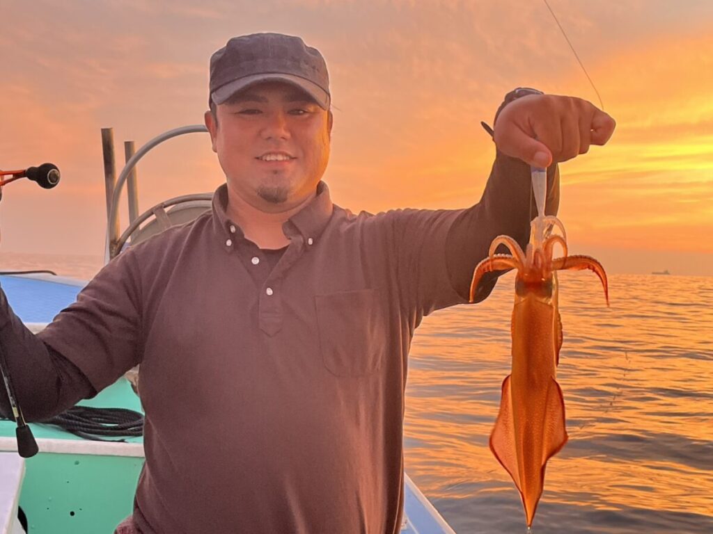 夕暮れ時、穏やかな海とオレンジ色の空を背景に、ボートに乗った笑顔の人が片手で獲れたてのイカを掲げている。.