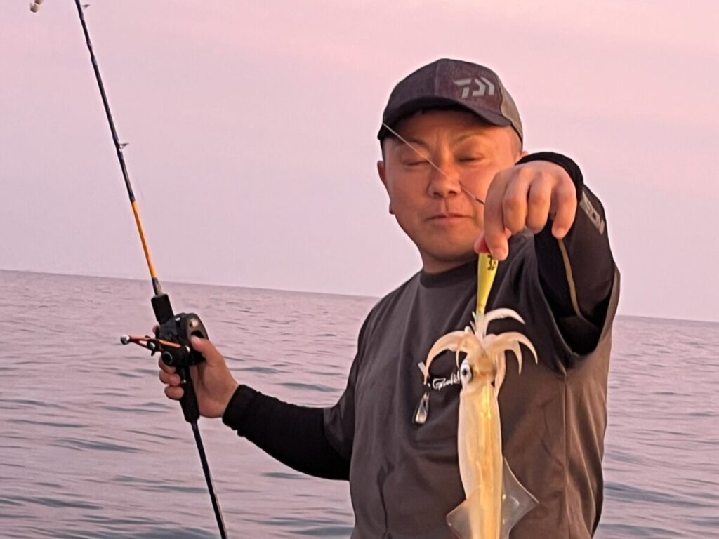 帽子をかぶり、釣り竿を持った人物が、獲れたてのイカを見せている。背景には海とピンクがかった空が広がっている。.