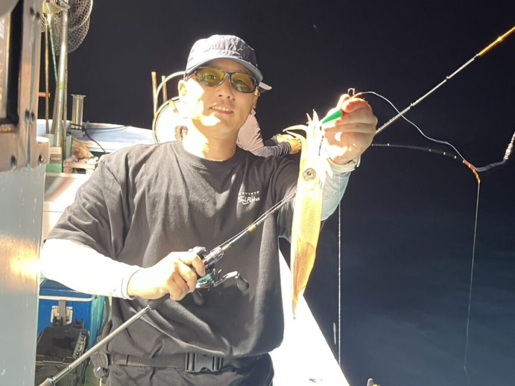 夜、ボートの上でサングラスとキャップをかぶった人物が、釣り竿を手に持ち、釣れたてのイカを見せながら笑っている。.