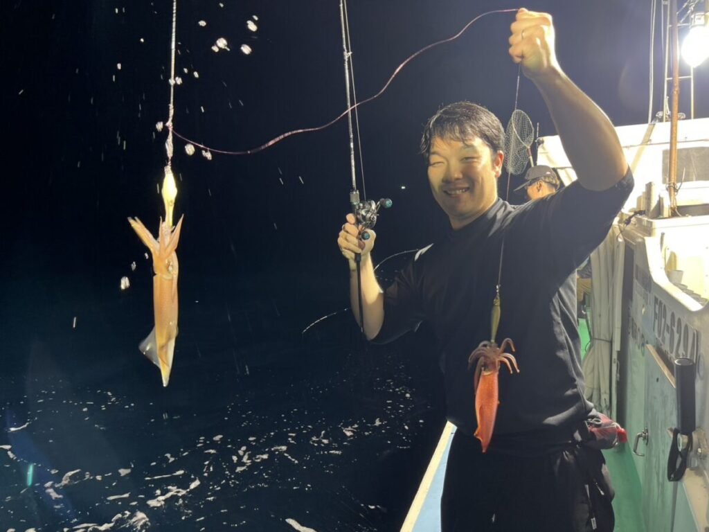 夜、ボートに乗った人物が笑顔で釣り竿を手にしており、別々の釣り糸にイカが2匹掛かっている。周囲には水しぶきが飛び散り、背景にはボートの装備が見える。.