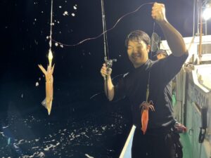 夜、ボートに乗った人物が笑顔で釣り竿を手にしており、別々の釣り糸にイカが2匹掛かっている。周囲には水しぶきが飛び散り、背景にはボートの装備が見える。.