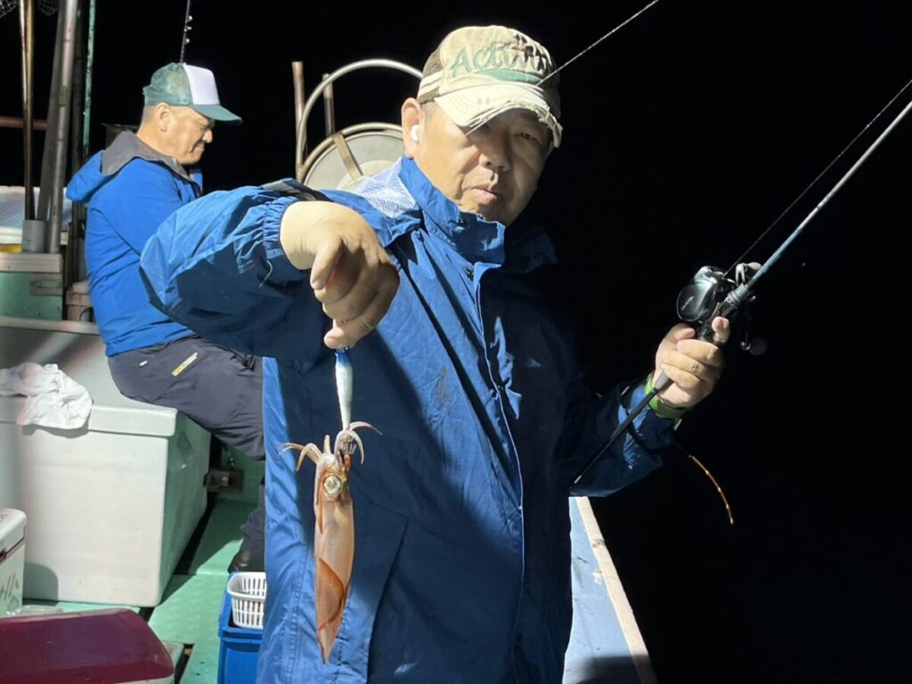 青いジャケットとキャップを身につけた男性が、夜、船上で釣りをしながら、釣り上げたばかりのイカを掲げている。背景には、同じく青いジャケットを着た別の人物が座って釣りをしている。.