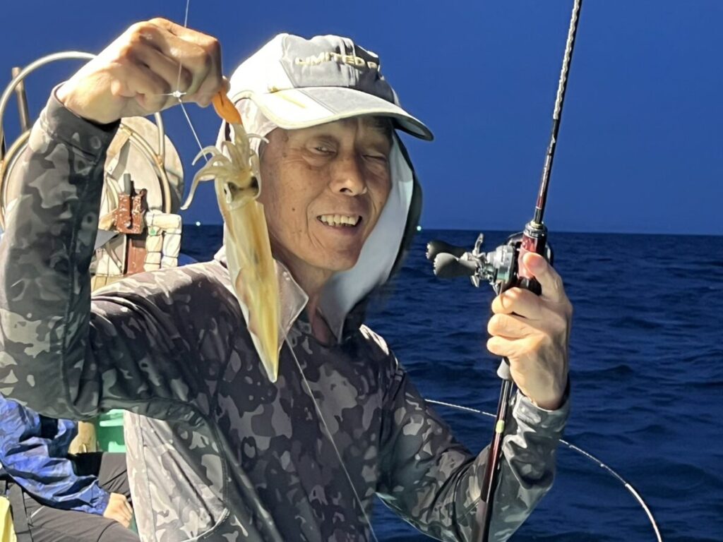 夕暮れ時の海の上、ボートの上に立つ男は、片手に釣り竿、もう片方の手には釣りたてのイカを持ち、キャップと柄物のシャツを着て微笑んでいる。.