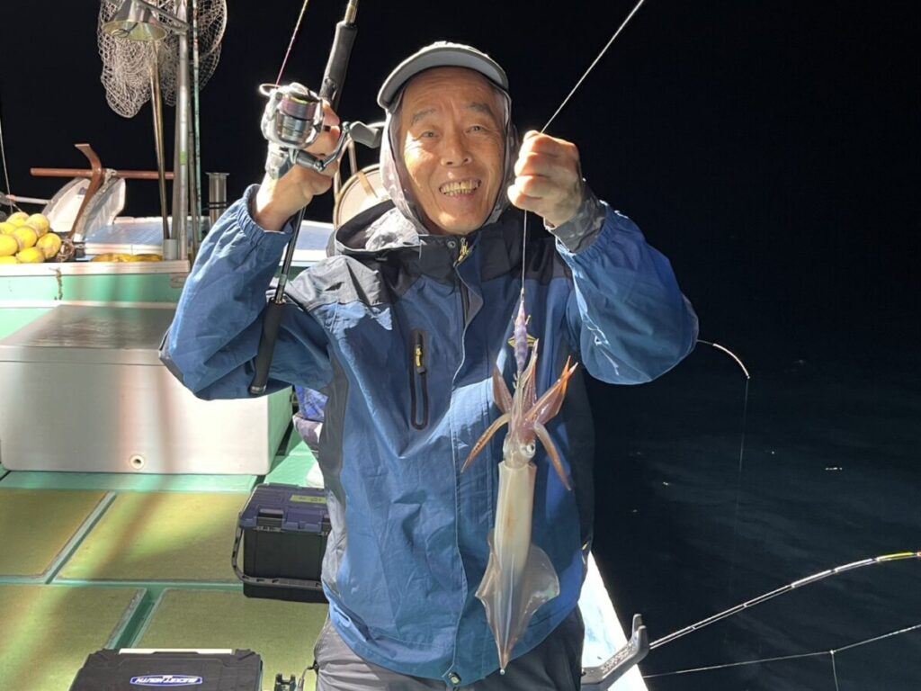 夜、ボートの上で、青いジャケットとキャップを身につけた笑顔の男性が、釣り竿を掲げている。竿には、釣り上げたばかりのイカが掛かっている。.