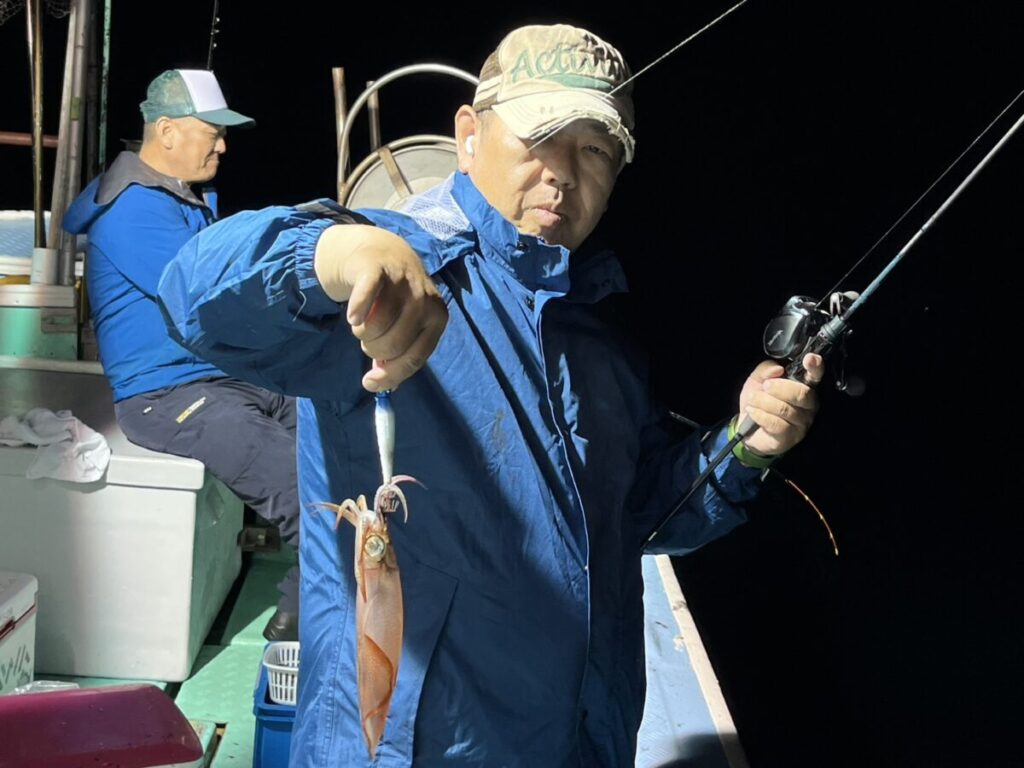 青いジャケットとキャップを身につけた男性が、夜、船上で釣りをしながら、釣り上げたばかりのイカを掲げている。その背後には、同じく青いジャケットを着た別の男性が座っている。暗い背景を背に、二人は明るい照明に照らされている。.