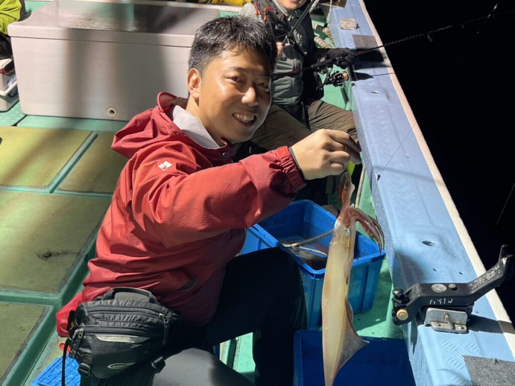 夜、赤いジャケットを着た男性がボートに座り、獲れたてのイカを掲げて微笑んでいる。背景には釣り道具と、もう一人の人物が見える。.