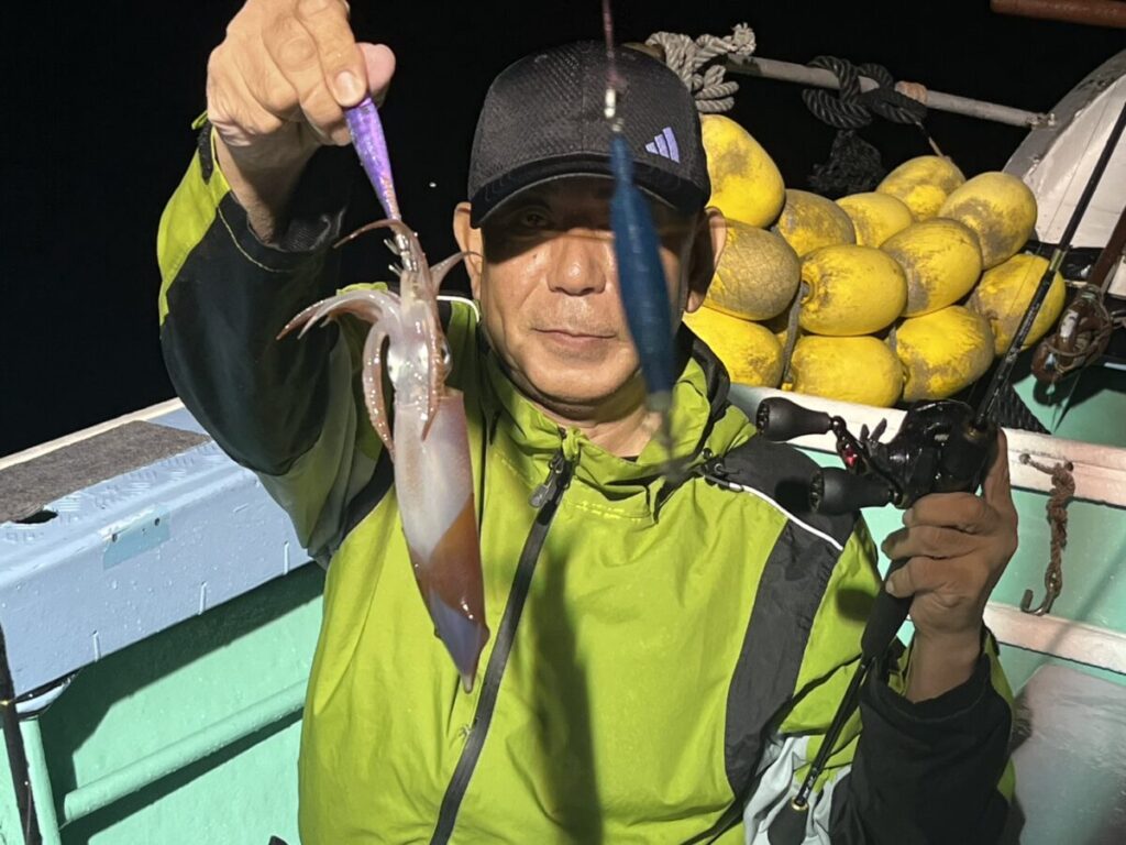 緑のジャケットと黒い帽子をかぶった人物が、夜間にボートで釣りをしながら、獲れたてのイカを掲げており、背景には黄色いブイが見える。.