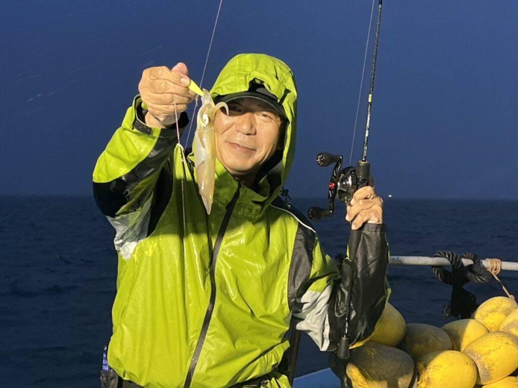 鮮やかな緑色のレインジャケットを着た人物が、釣り竿を手に、夜間にボートで釣った小さなイカを見せながら微笑んでいる。背景には海と黄色いブイがいくつか見える。.