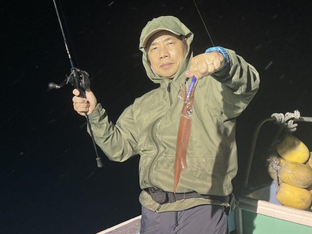 夜、ボートの上に立つ、緑色のレインジャケットとフードを身につけた人物が、片手に獲れたてのイカ、もう片方の手に釣り竿を掲げている。.
