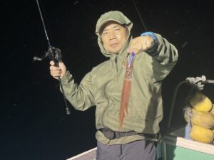夜、ボートの上に立つ、緑色のレインジャケットとフードを身につけた人物が、片手に獲れたてのイカ、もう片方の手に釣り竿を掲げている。.