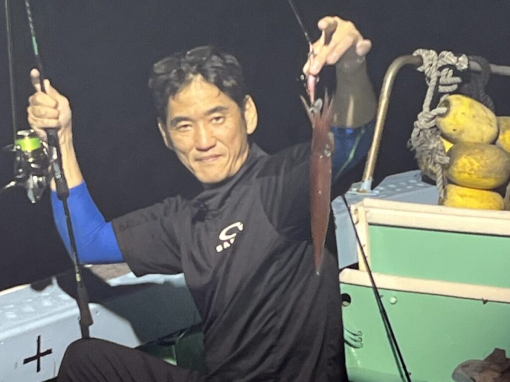 夜、ボートに乗った人物が、片手で釣り竿を掲げ、もう片方の手には獲れたてのイカを掲げ、カメラに向かって微笑んでいる。.
