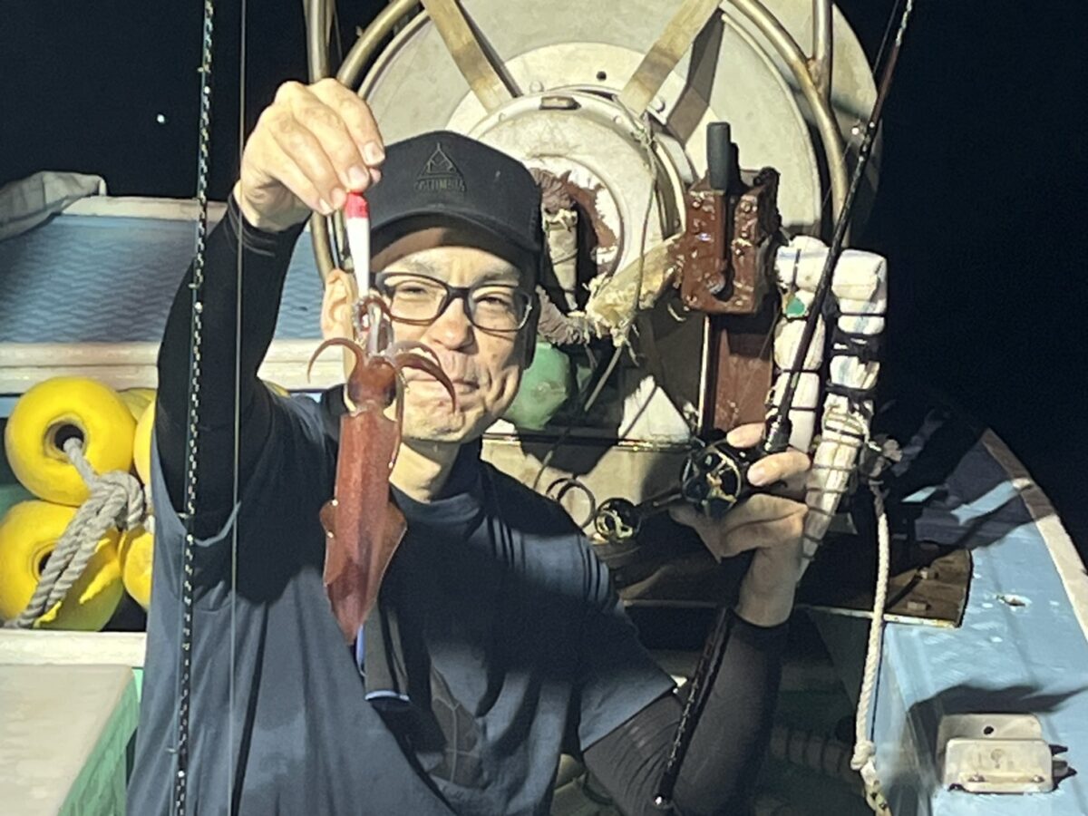 夜、漁船の上で、メガネと帽子をかぶった人物が小さなイカを掲げ、カメラに向かって微笑んでいる。背景にはさまざまな漁具が見える。.