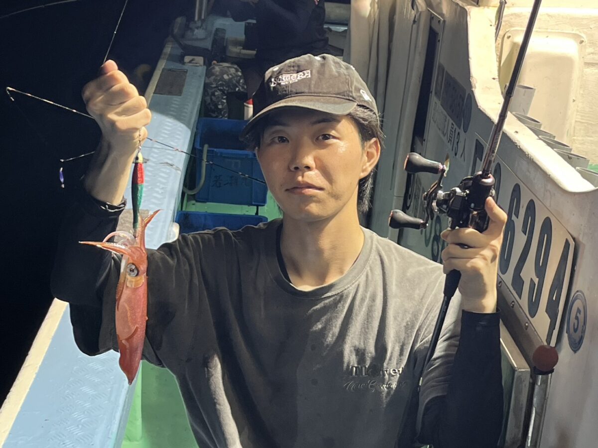 夜、ボートに乗った人物が、片手に釣り竿、もう片方の手に獲れたてのイカを持ち、カメラの方を見つめている。.