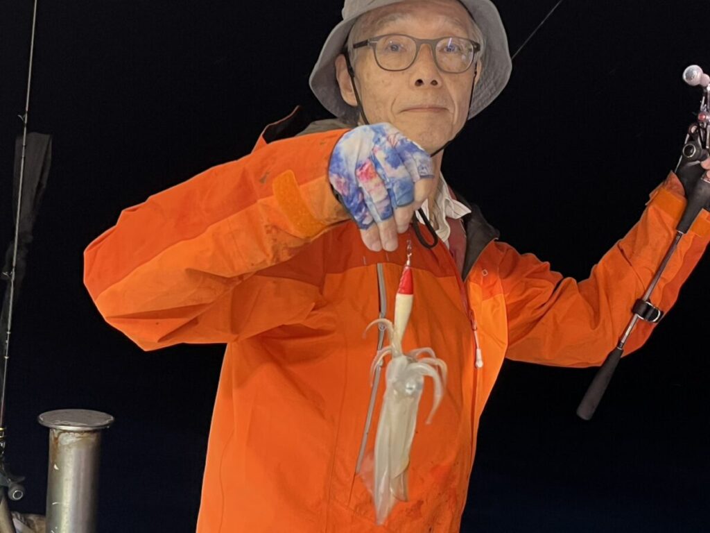 夜、オレンジ色のジャケットと灰色の帽子をかぶった人物が釣り竿を手にし、釣ったばかりのイカを見せている。.