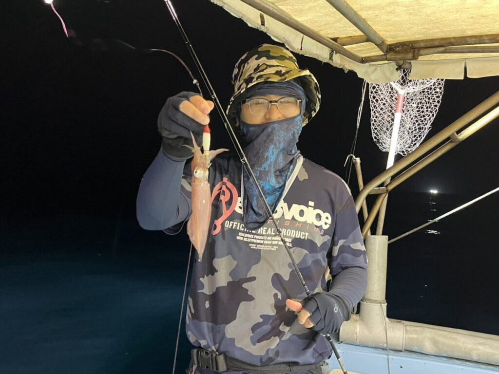 夜、ボートの上に立つ、迷彩服を着て顔を覆った人物が、釣り竿で釣ったイカを掲げている。背景には漁網が見える。.