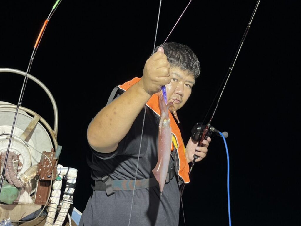 オレンジ色のライフジャケットを着た人物が、ボートでの夜釣りで釣った小魚を掲げており、背景には釣り竿や釣り道具が見える。.