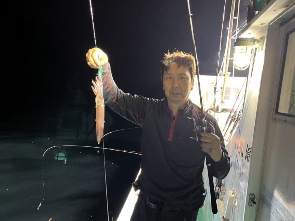 夜、ある人がボートの上に立ち、片手に釣り竿、もう片方の手には釣ったばかりのイカを持っている。ボートは明かりに照らされているが、背景は暗い。.