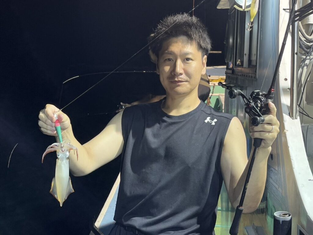 黒いノースリーブのシャツを着た男性が、夜、ボートに座り、片手に釣り竿、もう片方の手に釣ったばかりのイカを持っている。.
