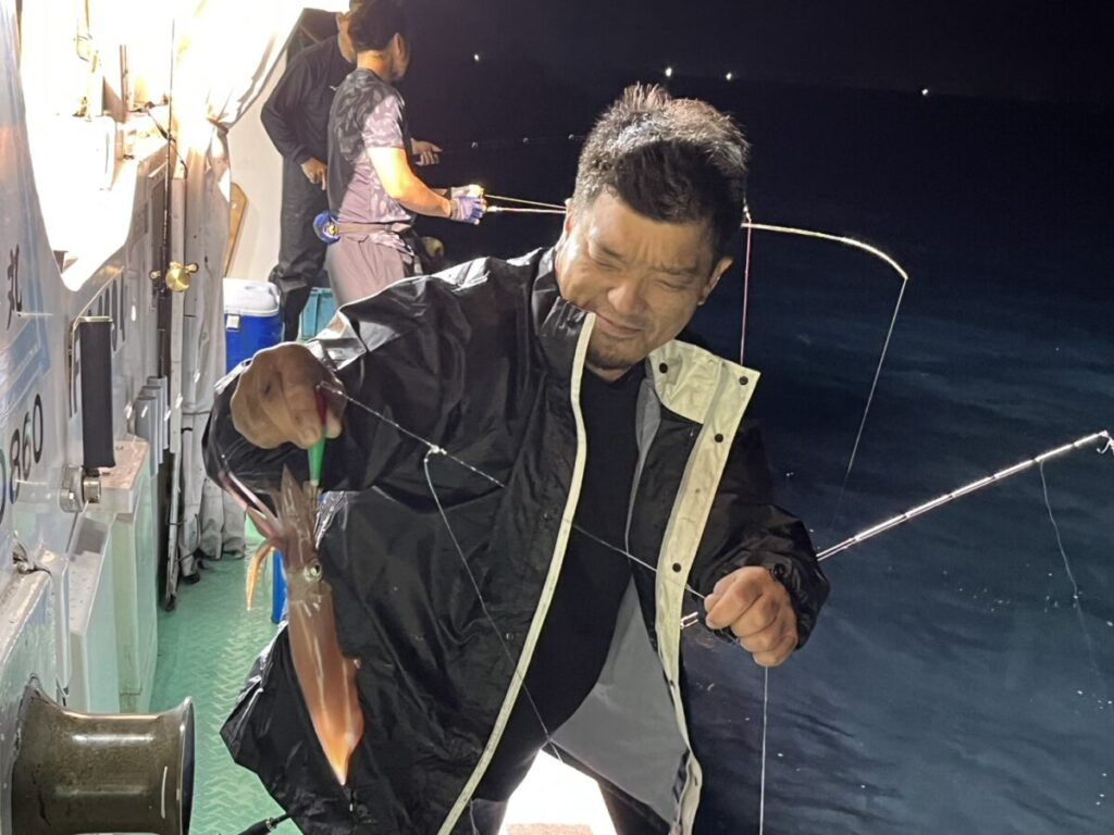 真っ黒なレインジャケットを着た男性が、釣り糸に掛かった獲れたてのイカを掲げて微笑んでいる。背景には深い青色の海が広がり、彼は夜の船の上に立っている。その近くでは、もう一人の人が釣りをしている。.