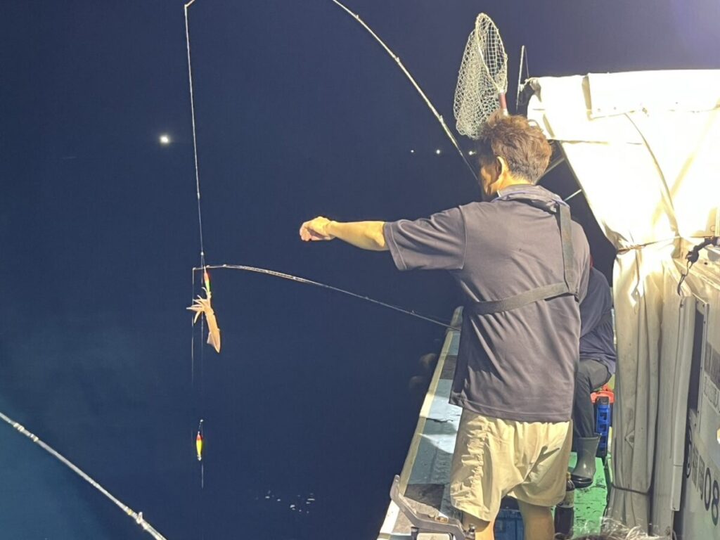 夜、ボートに乗った人物が釣りをしており、釣り糸にイカが掛かった竿を手にしている。ボートの上には釣り道具が見え、水面は暗く、わずかな光しか反射していない。.
