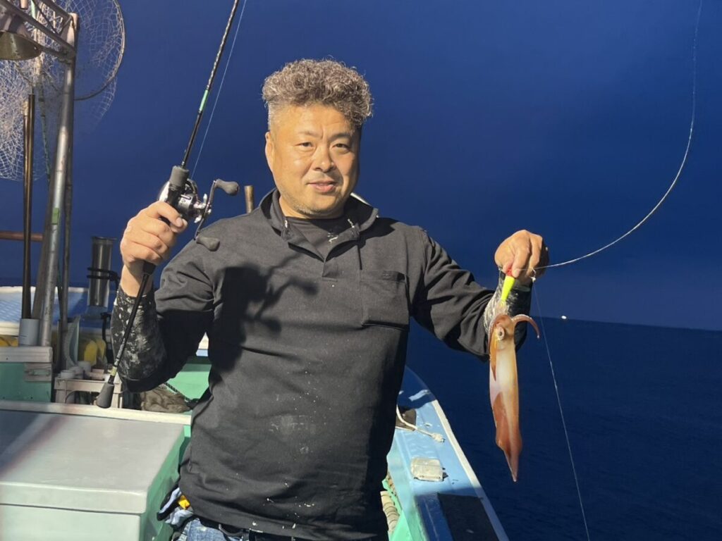 夜、ある男がボートの上に立ち、片手に釣り竿、もう片方の手には獲れたてのイカを持ち、背景には海と紺碧の空が広がっている。.