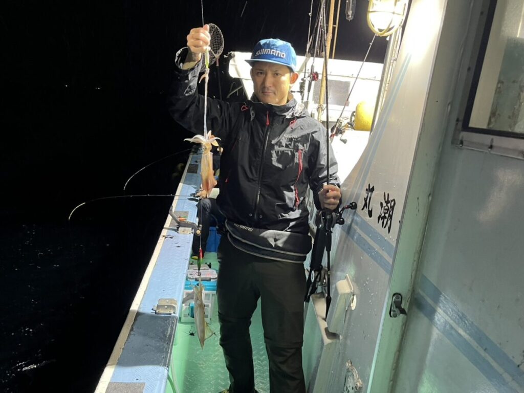 夜、ボートの上に立つ人物が、釣りで獲ったイカを掲げている。その人物は青いシマノのキャップをかぶり、レインウェアを着用している。デッキは濡れており、釣り道具が見える。.