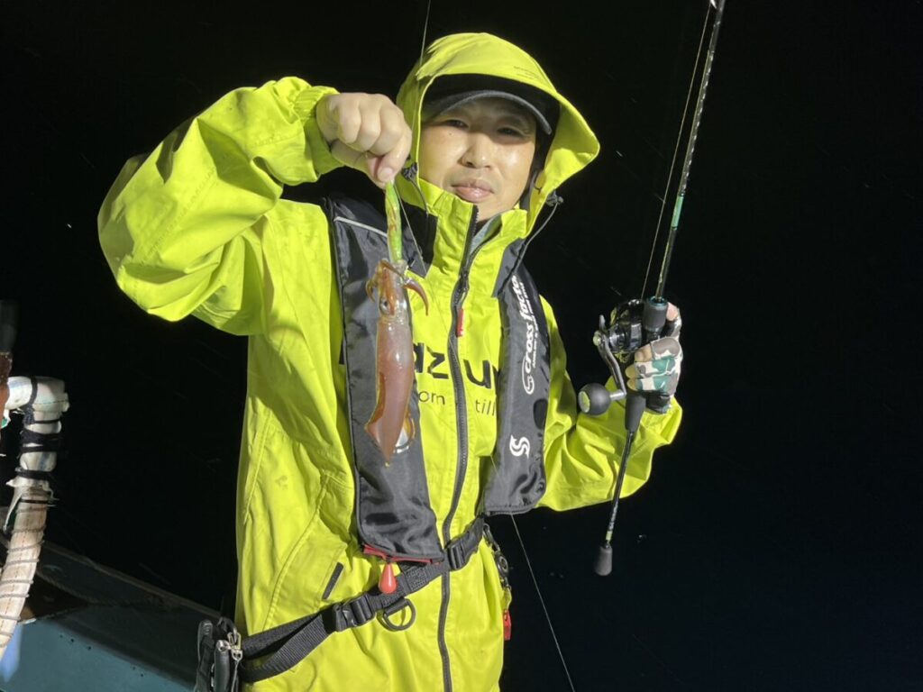 夜、ボートの上に立つ、鮮やかな黄色のレインジャケットと救命胴衣を着た人物が、片手に釣り竿、もう片方の手に獲れたてのイカを掲げている。.