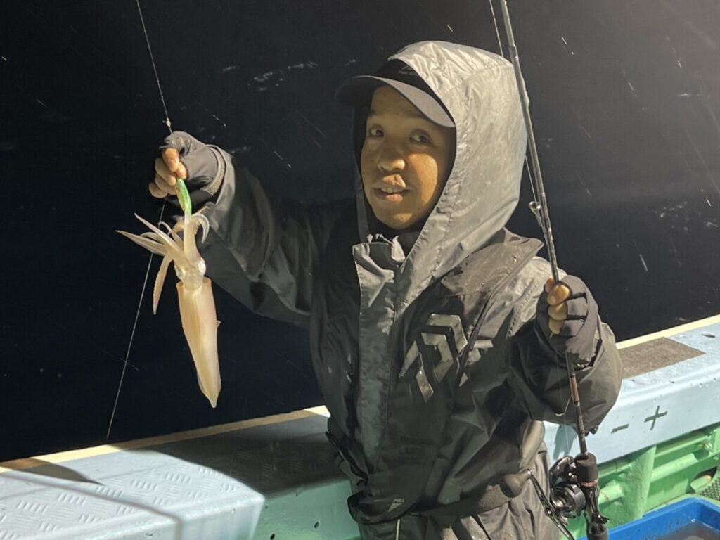 レインジャケットと帽子を身につけた子供が、夜釣りで釣ったイカを掲げている。その背後には、釣り道具が置かれたボートが見える。.