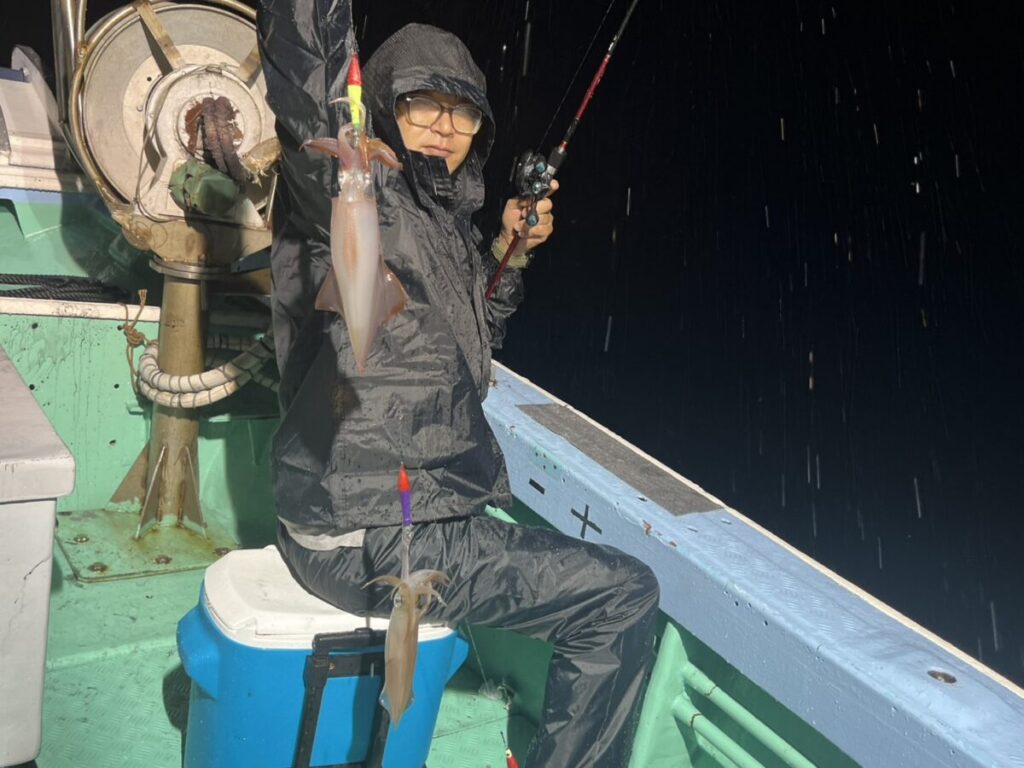 雨合羽を着て眼鏡をかけた人物が、夜、ボートの上に座り、別々の釣り糸にイカが2匹かかった釣り竿を手に持ち、雨が降っているのがはっきりとわかるにもかかわらず、カメラに向かって微笑んでいる。.