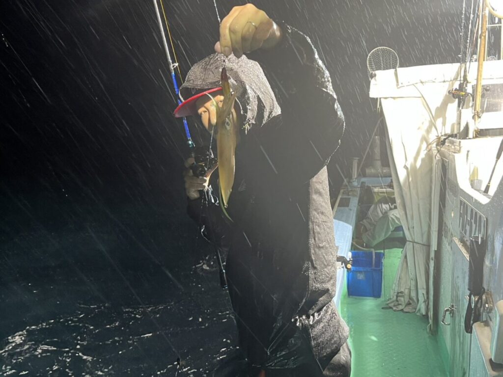 夜、雨の降る中、甲板が濡れて明かりに照らされた船の上で、フード付きのジャケットを着た人物が、釣り糸についたイカを掲げている。.