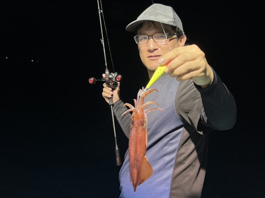 眼鏡と帽子をかぶった人物が、片手に釣り竿を持ち、もう一方の手で釣ったばかりのイカを見せている。暗い背景を背に立っており、おそらく夜のことだろう。.