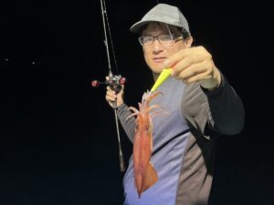 眼鏡と帽子をかぶった人物が、片手に釣り竿を持ち、もう一方の手で釣ったばかりのイカを見せている。暗い背景を背に立っており、おそらく夜のことだろう。.