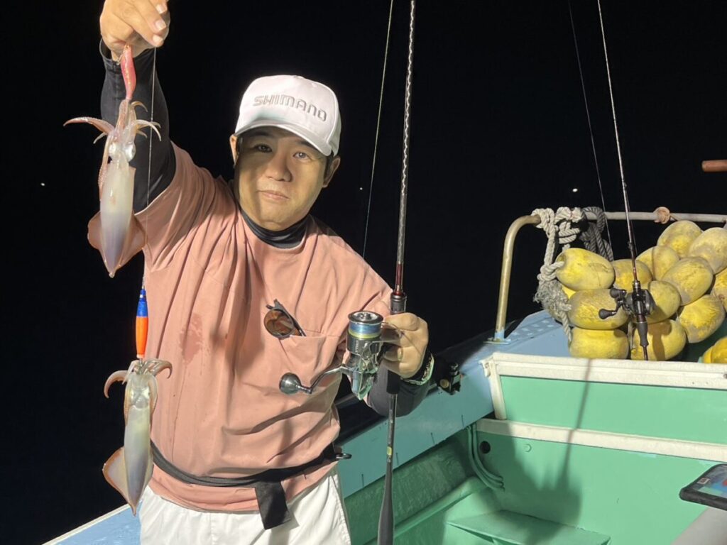 ボートに乗った人物が、片手に釣り竿、もう片方の手に獲れたてのイカ2匹を持ち、夜釣りをしている。背景には黄色いブイが見える。.