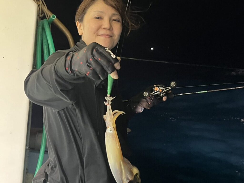 黒い手袋とジャケットを着た人物が、片手に釣り竿、もう片手に獲れたてのイカを持ちながら微笑んでいる。背景には夜の暗い水面と緑色のホースが見える。.