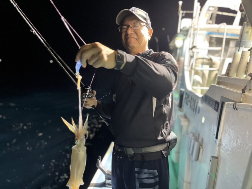 夜、ボートに乗った男性が、釣り竿で釣ったイカを手に持っている。彼は眼鏡をかけ、キャップをかぶり、濃い色のジャケットを着ている。ボートの明かりが scene を照らし出し、背景には海が見える。.