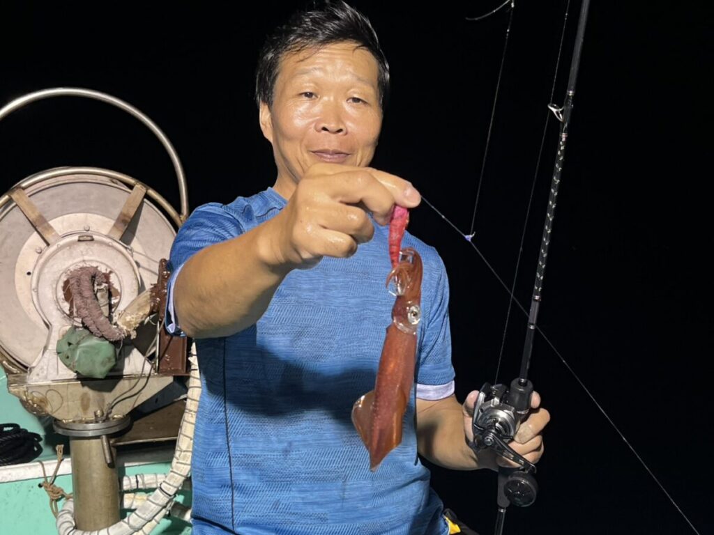 青いシャツを着た男性が、ボートでの夜釣りで釣った小さなイカを掲げ、カメラに向かって誇らしげに微笑んでいる。ボートの上には、彼の横に釣り道具やリールが見える。.