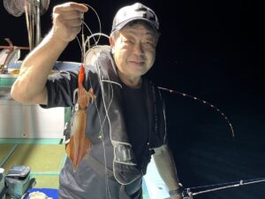 夜、ボートに乗った男が、釣り竿と釣り糸で釣ったばかりのイカを掲げながら微笑んでいる。.