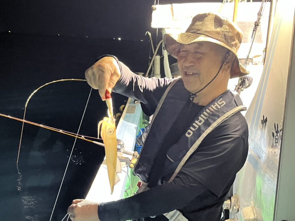 明るい明かりに照らされた夜の船上で、帽子をかぶった笑顔の人が、釣り道具を使ってイカを手にしている。.